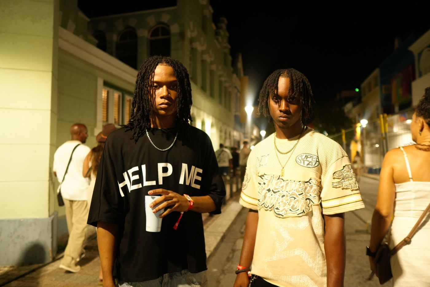 Two young men with locs standing on a lit street at night